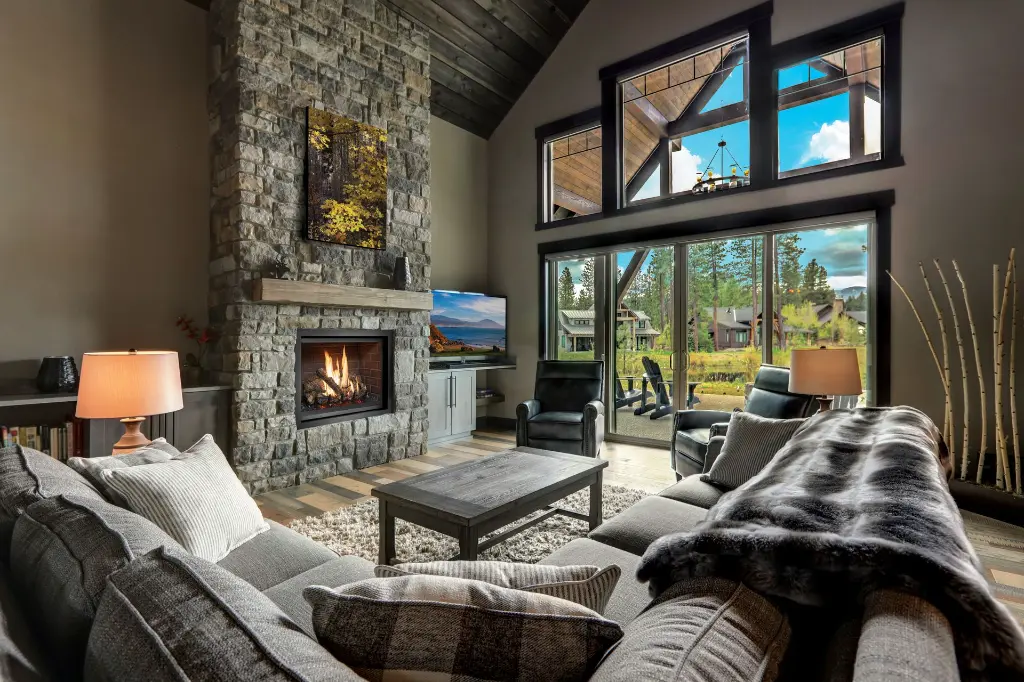 Ambiance "Modern Lodge" avec le foyer au gaz 864 TV 40K "Clean Face" dans une cheminée en pierres, avec des poutres de bois apparentes et de grandes fenêtres.