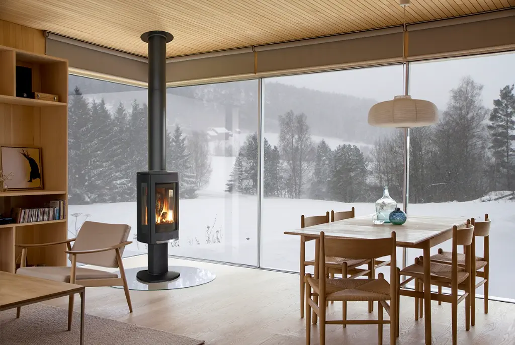 Poêle au gaz Jotul GF 370 noir mat sur piédestal, créant une ambiance chaleureuse dans un décor scandinave avec vue sur un paysage d'hiver.